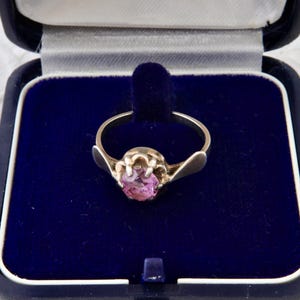 Può includere: Un anello d'argento con una pietra preziosa rosa, presentato in un cofanetto di velluto blu. L'anello presenta una pietra centrale di forma ovale e spalle decorative. Il cofanetto è aperto, rivelando l'anello.