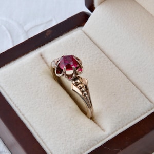 Puede incluir: Un anillo con una gema facetada de color rojo intenso y vibrante, engastada en una montura de metal de seis puntas. La banda está hecha de un material metálico y presenta un diseño decorativo y texturizado a lo largo de los hombros.