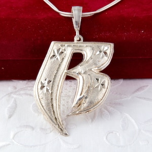 May include: A silver pendant necklace with a large, stylised letter "B". The pendant has a textured surface with starburst designs and hangs from a thin, silver chain. The background is a red velvet surface.
