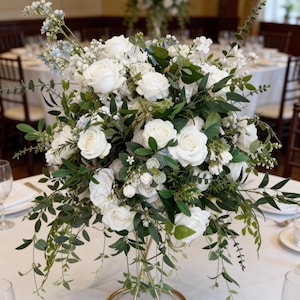 May include: A floral centerpiece featuring white roses, greenery, and delicate white flowers. The arrangement is displayed in a gold metal stand. The table setting includes white linens and glassware, creating an elegant ambiance.