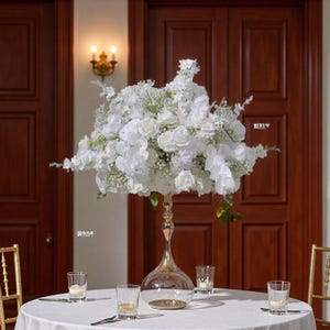 Könnte beinhalten: Ein hoher, eleganter Mittelstück mit einem großen Arrangement weißer Blumen, darunter Rosen und Orchideen. Das Blumendisplay steht auf einem goldfarbenen, sanduhrförmigen Ständer mit einer transparenten Glaskugelbasis. Mehrere kleine Gläser sind darum herum angeordnet.