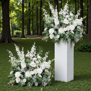 May include: Two displays of white flowers, including roses and delicate sprigs, are shown outdoors. One arrangement is on a white pedestal, the other is low to the ground. The background features a green lawn and a forest of tall trees.