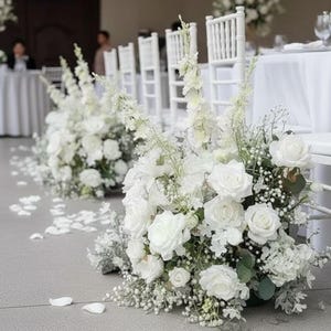 May include: A wedding ceremony setup with white floral arrangements and white chairs. The floral arrangements include roses and other white flowers. White petals are scattered on the floor. Tables are set with white tablecloths and glassware.