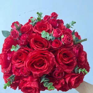 Op de afbeelding: Een levendig boeket rode rozen, met bloemen van verschillende groottes, geaccentueerd door kleine groene bladeren. De opstelling is dicht opeengepakt, waardoor een volle, ronde vorm ontstaat. De rozen hebben een diepe, rijke rode kleur.