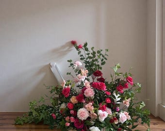 May include: A lush floral arrangement featuring a variety of roses in shades of red, pink, and white, with green foliage. The flowers are arranged on a wooden floor, leaning against a white wall. The arrangement includes a variety of rose sizes.