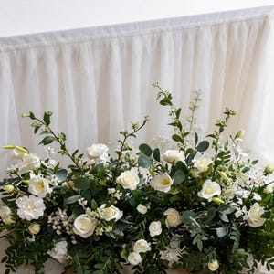 May include: A white table with a white tulle skirt and a floral arrangement. The arrangement features white roses, greenery, and other white flowers. The table is covered with a white tablecloth.