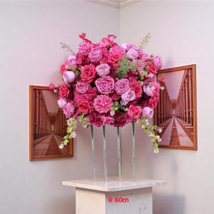 May include: A large, vibrant floral arrangement in shades of pink and magenta, supported by clear, slender stems. The arrangement is displayed on a white marble-like pedestal. The arrangement is approximately 90cm tall. Two framed pictures of a hallway are on the wall.