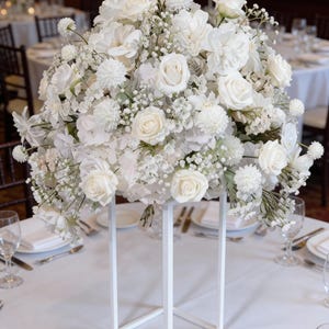 May include: A large, all-white floral display, including roses and hydrangeas, on a white geometric stand. The arrangement is set on a table, likely for a formal event. The table is set with white linens and glassware.