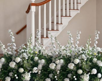 Op de afbeelding: Een bloemstuk met witte rozen en andere witte bloemen met groene bladeren. De opstelling staat voor een trap met een houten leuning en witte balusters. De vloer is van hout.