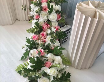 Puede incluir: Una larga guirnalda de rosas artificiales blancas y rosas con hojas verdes. La guirnalda está drapeada sobre una superficie blanca y se utiliza como decoración de boda o evento.