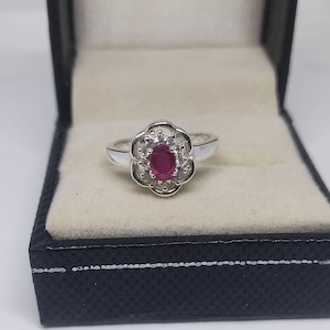 May include: A silver ring with a flower-shaped setting. The center features an oval, deep red gemstone, surrounded by smaller, clear stones. The ring is presented in a black jewelry box with a cream-colored interior.