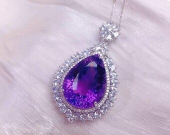 May include: A silver necklace featuring a large, pear-shaped purple amethyst pendant. The amethyst is surrounded by a halo of smaller, clear gemstones. The pendant hangs from a delicate silver chain, creating an elegant jewelry piece.