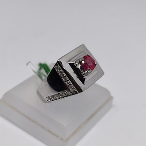 May include: A silver ring with a rectangular design, featuring a vibrant red gemstone at the top. The ring is accented with a row of small, clear stones along one side. The ring is displayed on a white stand.
