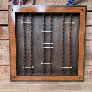 May include: A wooden display case with a dark brown interior and a lighter brown frame. The case has vertical slots and white pegs for holding items. The frame is secured with black screws. Decorative fabric hangs above the case.