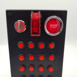 Könnte beinhalten: Schwarzes Panel mit 16 roten Druckknöpfen, 4 schwarzen Knöpfen, einem roten Kippschalter, einem silbernen Schlüssel und einem roten Knopf mit dem Text "MOTOR START" in weiß.