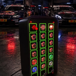 May include: A black rectangular control panel featuring a grid of illuminated buttons and switches with green borders. The panel includes a prominent red engine start button, various circular toggle switches, and rotary knobs, all set against a carbon fiber textured background.