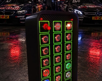 Puede incluir: Un panel de control rectangular negro con una cuadrícula de botones e interruptores iluminados con bordes verdes. El panel incluye un botón rojo de arranque de motor, varios interruptores circulares y perillas giratorias sobre un fondo con textura de fibra de carbono.