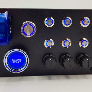 Könnte beinhalten: Eine schwarze Platte mit einem blauen Kippschalter, sieben blauen beleuchteten Druckknöpfen, drei schwarzen Knöpfen und einem großen blauen beleuchteten Druckknopf mit der Aufschrift "MOTOR START".