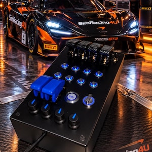 May include: A black sim racing button box featuring four top-mounted carbon fibre toggle switches, two large blue safety-covered switches, a central engine start button, multiple illuminated blue push buttons, and four rotary dials with blue indicators.