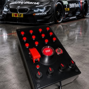 Può includere: Un pannello di controllo rettangolare nero dotato di numerosi pulsanti rossi, un grande pulsante centrale rosso con la scritta ENGINE START, un interruttore di sicurezza rosso e tre manopole rotanti nere con indicatori rossi.