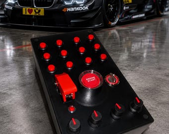 Puede incluir: Un panel de control rectangular negro con numerosos botones pulsadores rojos, un botón central grande rojo con la etiqueta ENGINE START, un interruptor de seguridad rojo y tres perillas giratorias negras con indicadores rojos.
