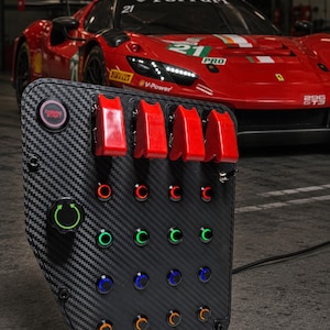 Op de afbeelding: Een koolstofvezel schakelpaneel met een rode Engine Start-knop, vier rode tuimelschakelaars en een raster van zestien verlichte drukknoppen in rood, groen, blauw en oranje. Een zwarte draaiknop met een groen cirkelvormig pijlpictogram is inbegrepen.