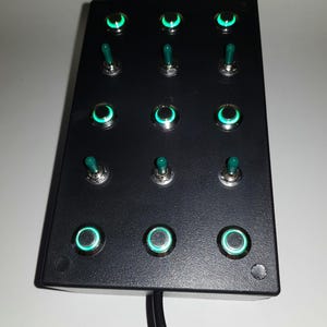 May include: A black box with twelve round, green-lit toggle switches. Each switch has a green button and a silver ring around the light. The box has a black cord attached to it.