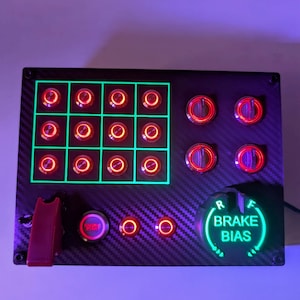 May include: A black panel with a grid of 12 red illuminated buttons, four larger red illuminated buttons, a red illuminated button labeled "Engine Start", and a green illuminated knob labeled "Brake Bias".