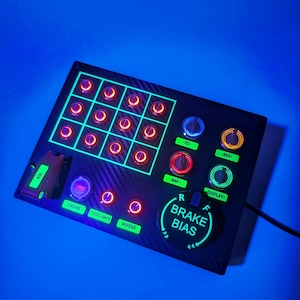 May include: A black panel with a carbon fiber texture and glowing green and red buttons. The panel has a variety of controls, including a brake bias knob, a display button, and a map button. The panel also has a series of toggle switches, including an ignition switch, an engine switch, and a cycle lights switch.