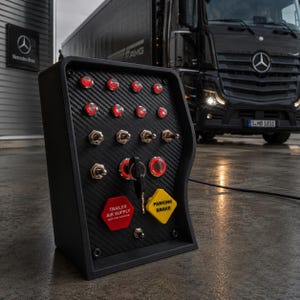 May include: A black control panel with a carbon fibre texture featuring multiple illuminated red push buttons, toggle switches, a central ignition key, and two distinct knobs labelled TRAILER AIR SUPPLY and PARKING BRAKE in bold text.