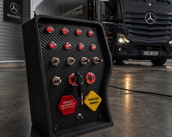 May include: A black control panel with a carbon fiber texture featuring multiple illuminated red push buttons, toggle switches, a central ignition key, and two distinct knobs labeled TRAILER AIR SUPPLY and PARKING BRAKE in bold text.