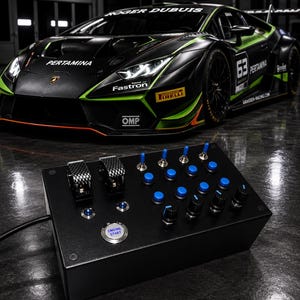 May include: A black rectangular control box featuring two carbon fibre toggle switches, several blue push buttons, black rotary knobs, and a silver Engine Start button. The device is positioned in front of a black and green Lamborghini race car.
