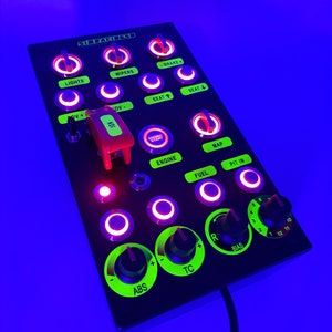 Könnte beinhalten: Ein schwarzes Bedienfeld mit grün und rot beleuchteten Knöpfen und Drehknöpfen. Das Bedienfeld ist mit "Lights", "Wipers", "ABS", "TC", "Bias", "Engine", "Fuel", "Map", "Pit In", "Seat", "-Brake", "OV", "IGN" und "SIRAGIC" beschriftet.