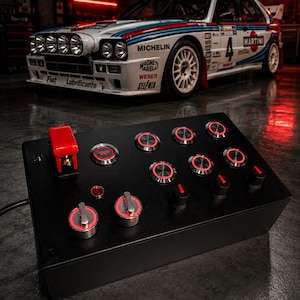May include: A black rectangular control panel featuring a red flip-up toggle switch, multiple circular push buttons with illuminated red rings, and several black rotary knobs. The device is positioned in front of a white Lancia rally car.