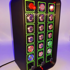 May include: A black control panel with a carbon fiber pattern and multiple buttons and knobs. The buttons are green, red, and blue. The panel has a red "Engine Start" button.