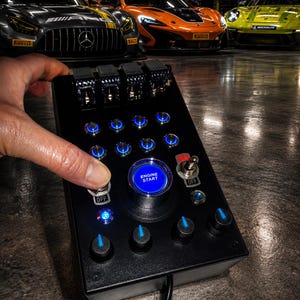 May include: A black rectangular button box controller featuring a large central blue illuminated button labeled ENGINE START, multiple smaller blue LED push buttons, four top-mounted toggle switches, and four rotary dials with blue indicators.