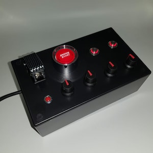 Peut inclure: Boîte noire avec un bouton rouge marqué "Démarrage du moteur" et plusieurs autres boutons et boutons rouges.