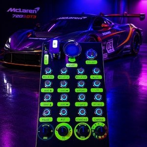 May include: A black racing simulator button box with illuminated green labels and blue-lit rotary encoders. The panel features switches for ignition, engine map, and various car settings, positioned in front of a McLaren 720S GT3 race car.