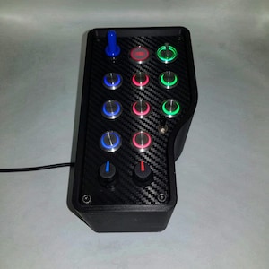Puede incluir: Un mando de juego negro con un diseño de fibra de carbono. Tiene 12 botones con luces azules, rojas y verdes. Hay un joystick azul en la parte superior y dos botones negros en la parte inferior.