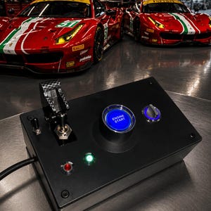 May include: A black rectangular control box featuring a prominent blue illuminated push button labelled ENGINE START, a carbon fibre toggle switch, a rotary dial, and two smaller indicator lights in red and green, with two red race cars in the background.