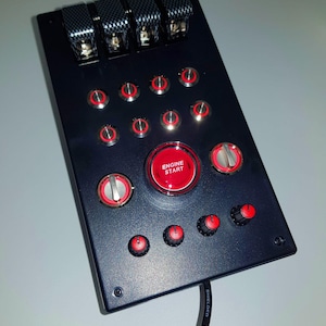 Puede incluir: Un panel negro con una variedad de botones rojos y plateados, incluyendo un botón rojo grande con la etiqueta "Arranque del motor". El panel tiene una textura de fibra de carbono negra en la parte superior y un cable negro conectado en la parte inferior.