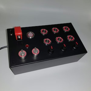 May include: A black box with a red toggle switch and multiple round buttons with red LED lights. The buttons are arranged in rows and columns. There are also two round knobs with red accents.