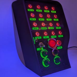 Op de afbeelding: Een zwart paneel met een verscheidenheid aan rood en groen verlichte knoppen en schakelaars. Het paneel heeft een carbonfiberpatroon en bevat labels zoals "Stoel", "Flits", "Ruitenwissers", "Motec", "Blackbox", "Tear Off", "Info", "Camera", "Straal", "Display", "Contact", "Pit Red", "100", "200" en "Boost".