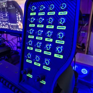 May include: A black control panel with numerous illuminated buttons and switches, labeled with text like "Battery Power" and "Eng R Start." The panel has a carbon fiber texture and is lit with blue and green lights, suggesting a cockpit or simulator setup.