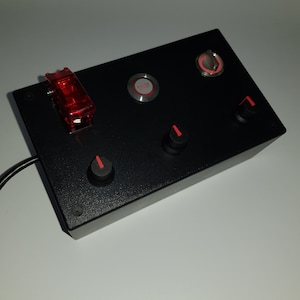 Puede incluir: Panel de control negro con tres botones rojos, dos perillas y un interruptor de palanca rojo. Los botones están etiquetados como "Arranque del motor" y "Turbo".