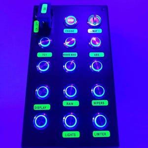 Può includere: Un pannello nero con una varietà di pulsanti illuminati, tra cui "Motore", "TC1", "Freno a mano", "ABS", "Pioggia", "Tergicristalli", "Display", "Luci" e "Limitatore". Il pannello presenta anche un pulsante rosso "Avvio" e una manopola etichettata come "Mappa". Il pannello è etichettato come "PS5" nella parte inferiore.