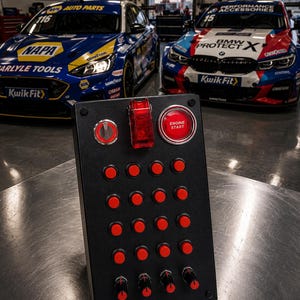 Può includere: Un pannello di controllo rettangolare nero con un interruttore a levetta rosso, un pulsante circolare rosso con la scritta ENGINE START, un piccolo interruttore argentato, sedici pulsanti rossi in griglia e quattro manopole nere rotanti.