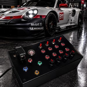 May include: A black rectangular button box controller featuring multiple red toggle switches, illuminated push buttons in various colours, and rotary knobs. A carbon fibre style safety cover protects the engine start button on the left side.