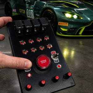 May include: A black rectangular button box controller featuring four carbon fiber toggle switches at the top, a large central red Engine Start button, a metal On-Off toggle switch, multiple small illuminated red push buttons, and four rotary dials.