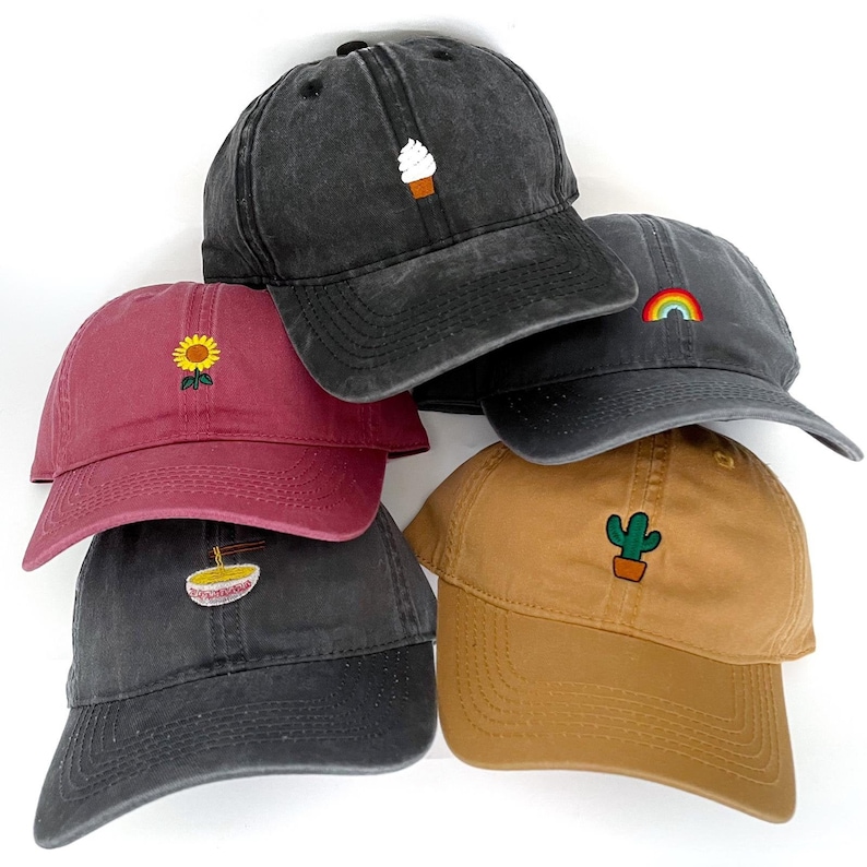 Puede incluir: Cinco gorras de b&eacute;isbol de diferentes colores con dise&ntilde;os bordados. Las gorras son negras, granate, marrones, negras y tostadas. Los dise&ntilde;os son un cono de helado, un girasol, un arco&iacute;ris, un cuenco de fideos ramen y un cactus.